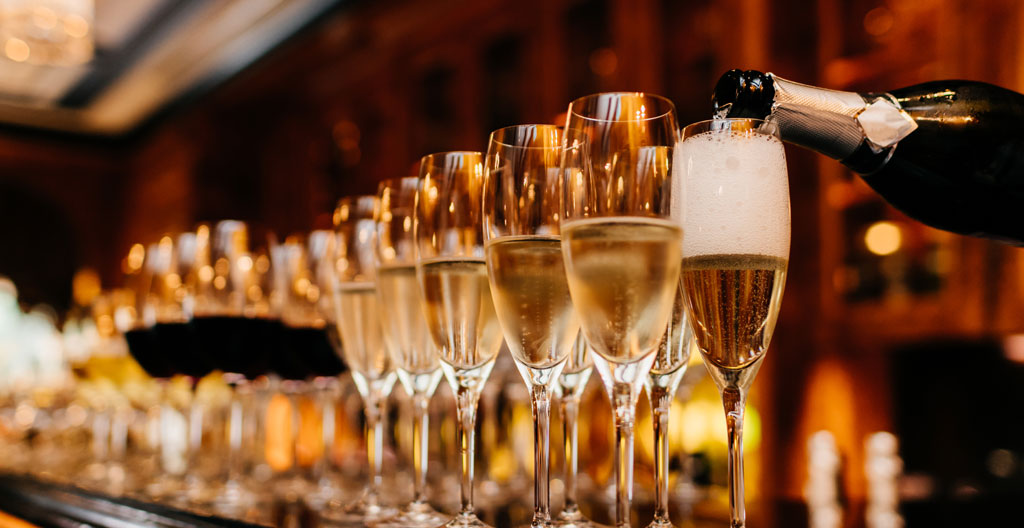 Main versant du champagne dans un verre tulipe incliné à 45 degrés sur un comptoir en pierre claire, mousse se formant délicatement